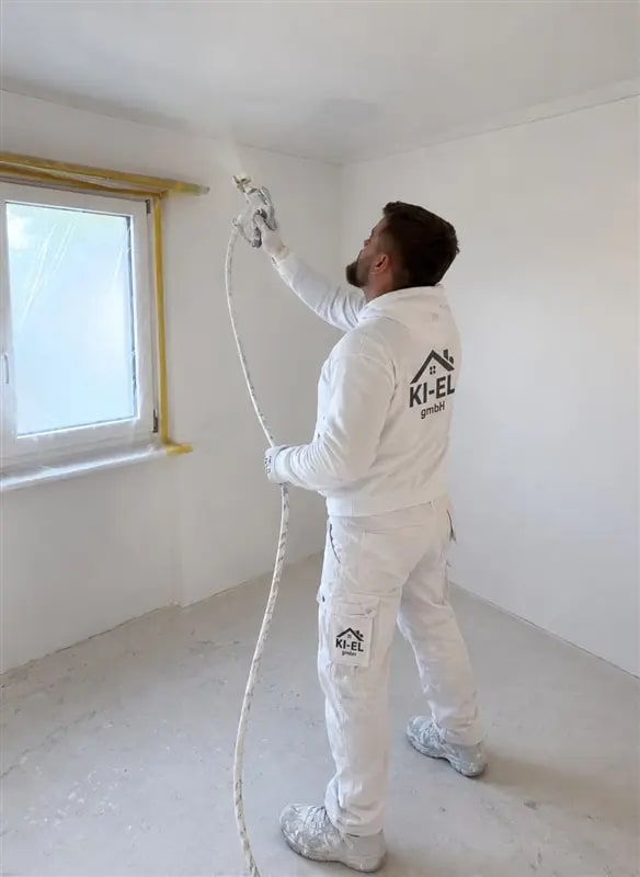 Mitarbeiter in KI-EL-Arbeitskleidung arbeitet mit einem Spritzgerät an einer Wand in einem leeren Raum, das Fenster ist abgeklebt