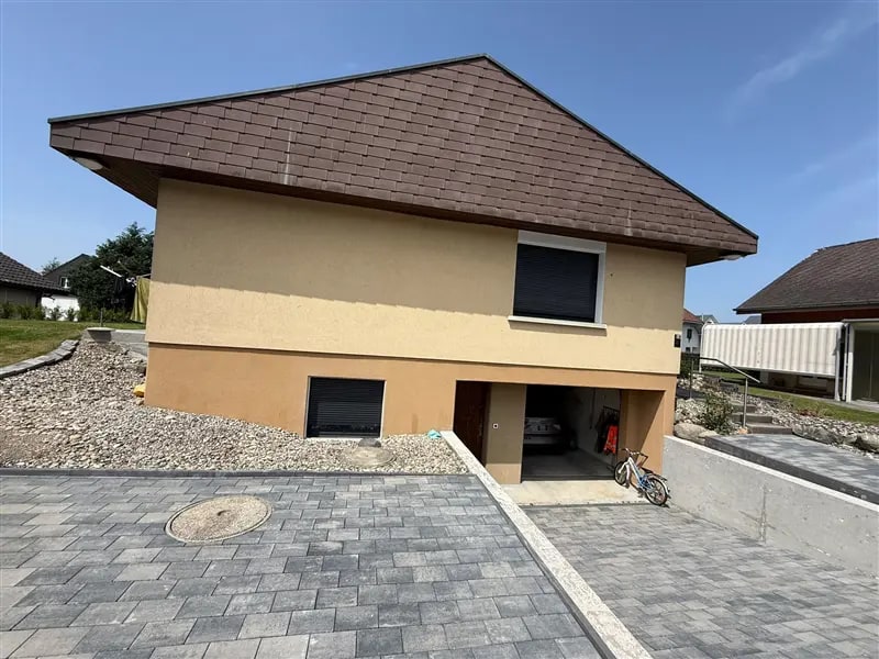 Einfamilienhaus mit gelber Fassade und braunem Dach, davor die gepflasterte Einfahrt